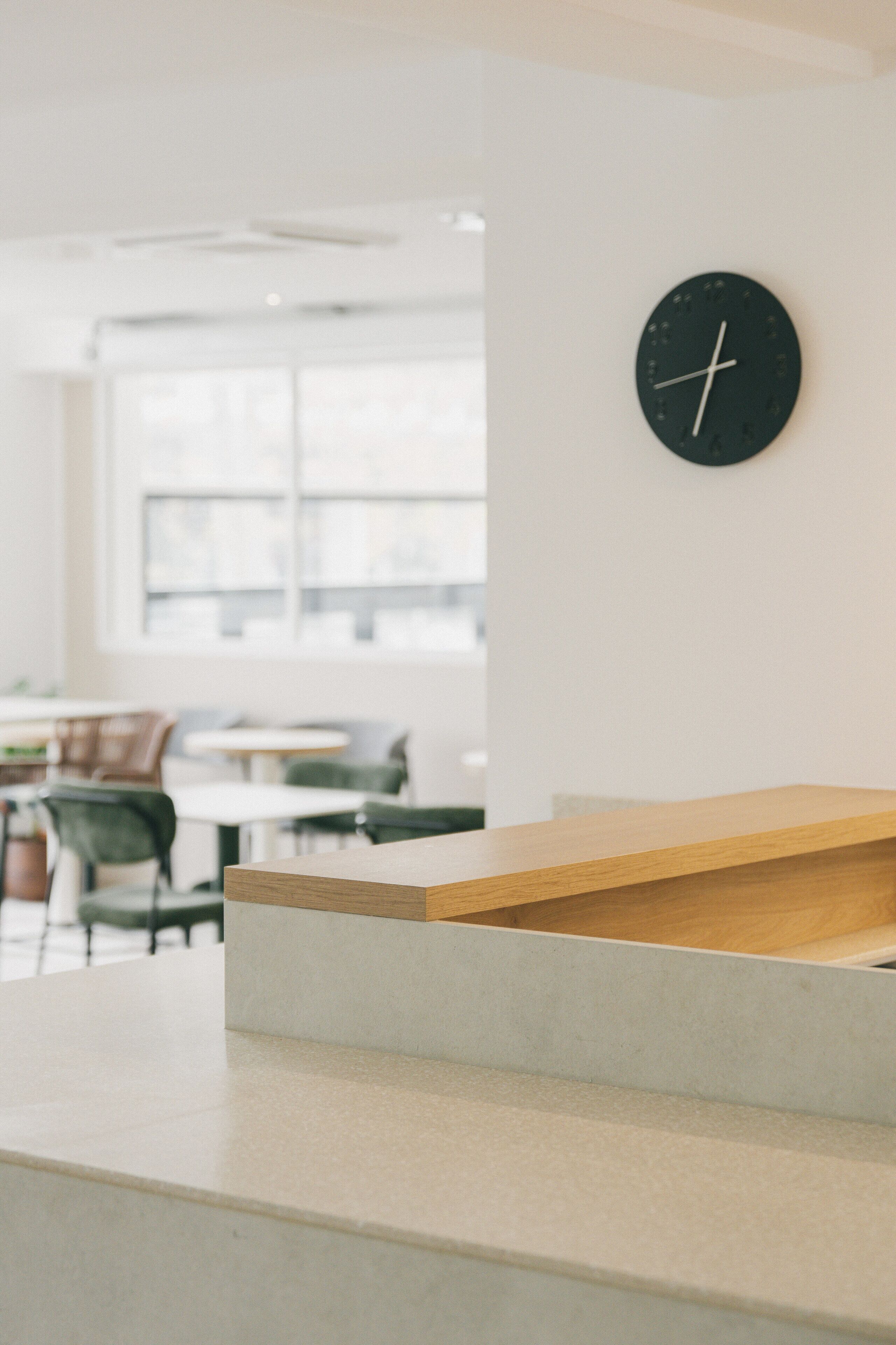 A modern interior with a wooden bench, a black clock on the wall, and a view of a dining area with tables and chairs.