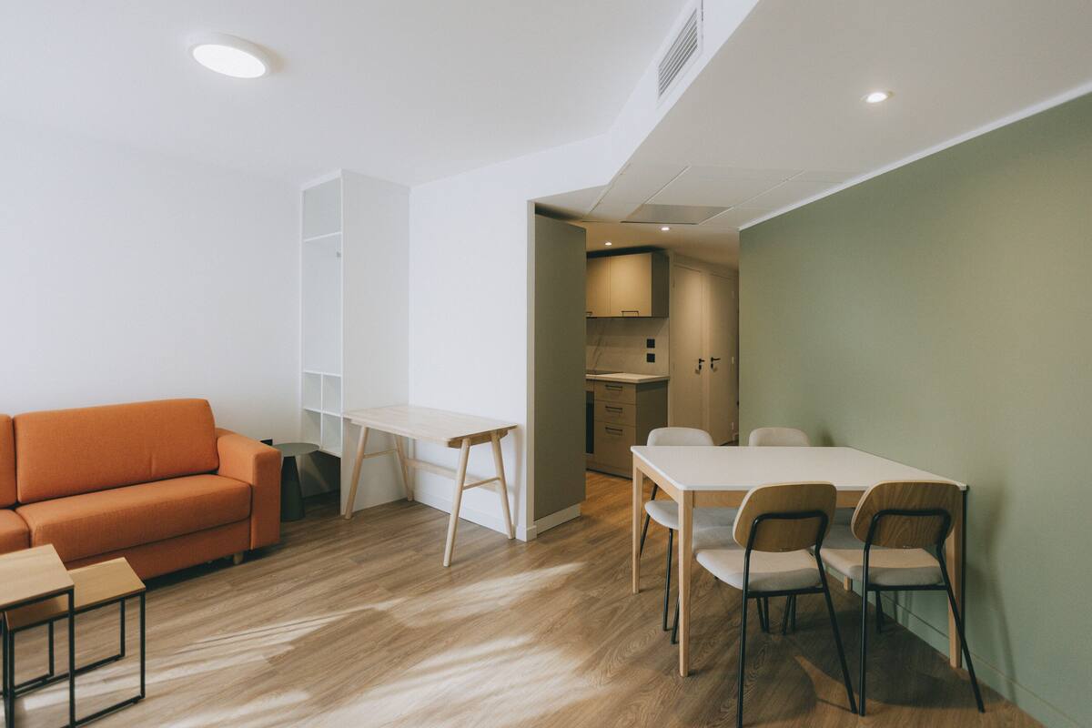 Un salon moderne avec un canapé, une table à manger et des chaises, et un coin cuisine visible à travers une porte ouverte.