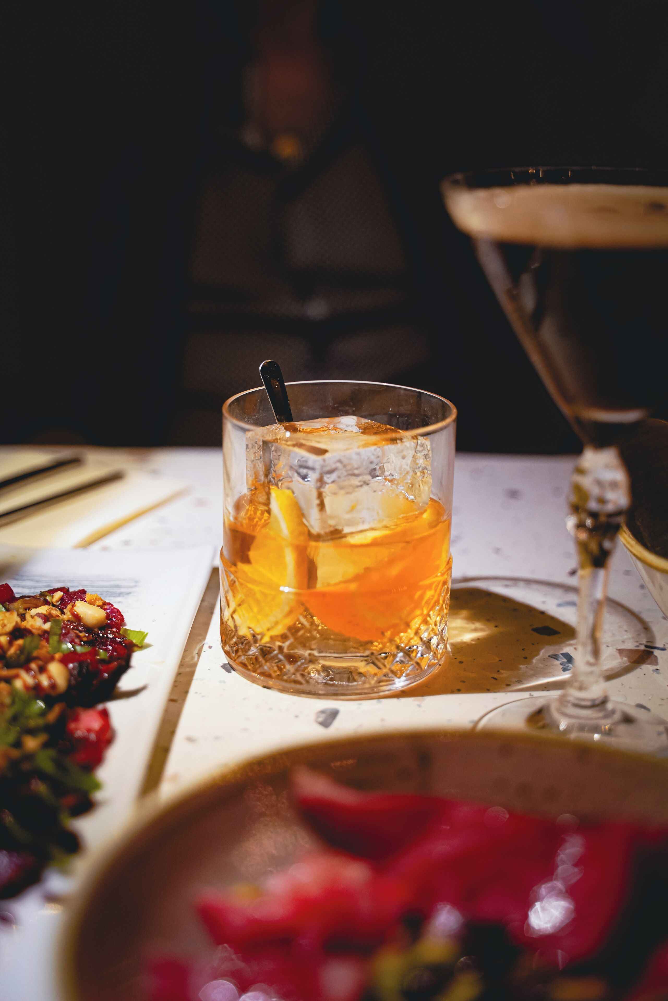 Un verre d’une boisson de couleur ambrée, contenant des glaçons et une tranche d’orange, posé sur une table avec une serviette et une fourchette à gauche.