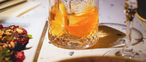 A glass of amber-colored beverage with ice cubes and a slice of orange, placed on a table with a napkin and a fork to the left.