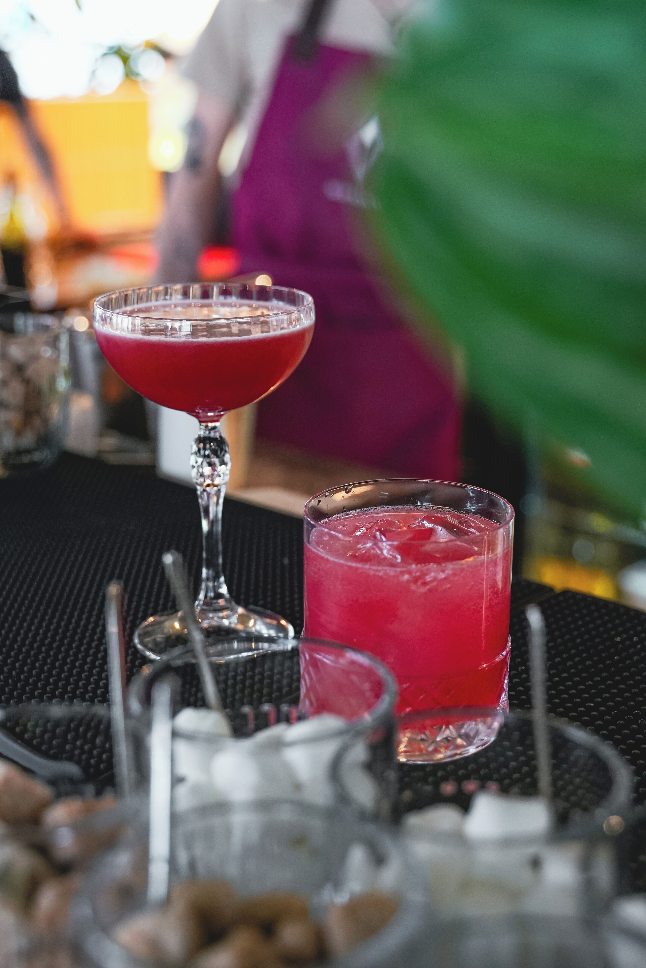 A bar setting with two glasses of red beverage, a cocktail shaker, and a bottle on a black textured surface.