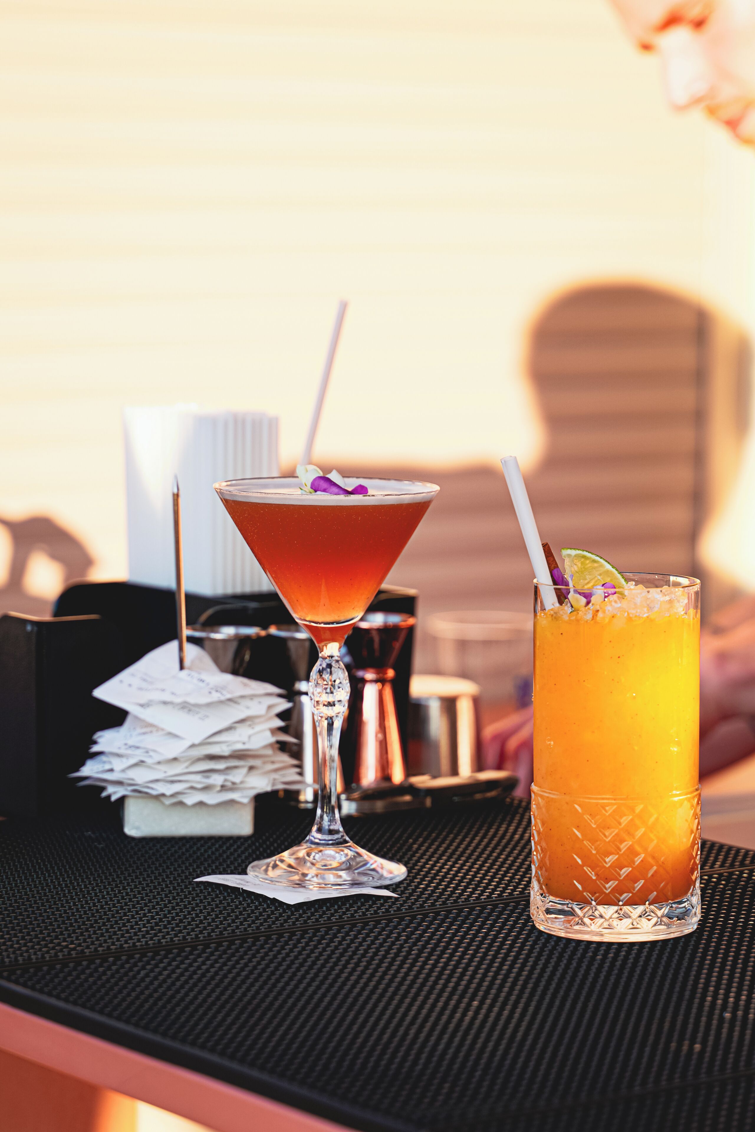 Twee cocktails op een tafel met een zwarte onderzetter, een stapel witte servetten en een metalen bak.