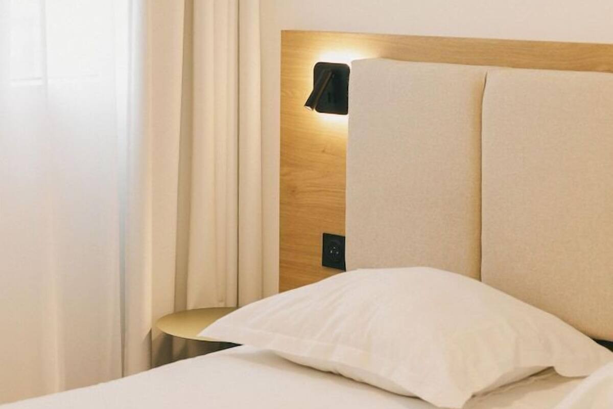 Une chambre d’hôtel avec un lit, une table de chevet, une lampe fixée au mur et une fenêtre avec des rideaux.