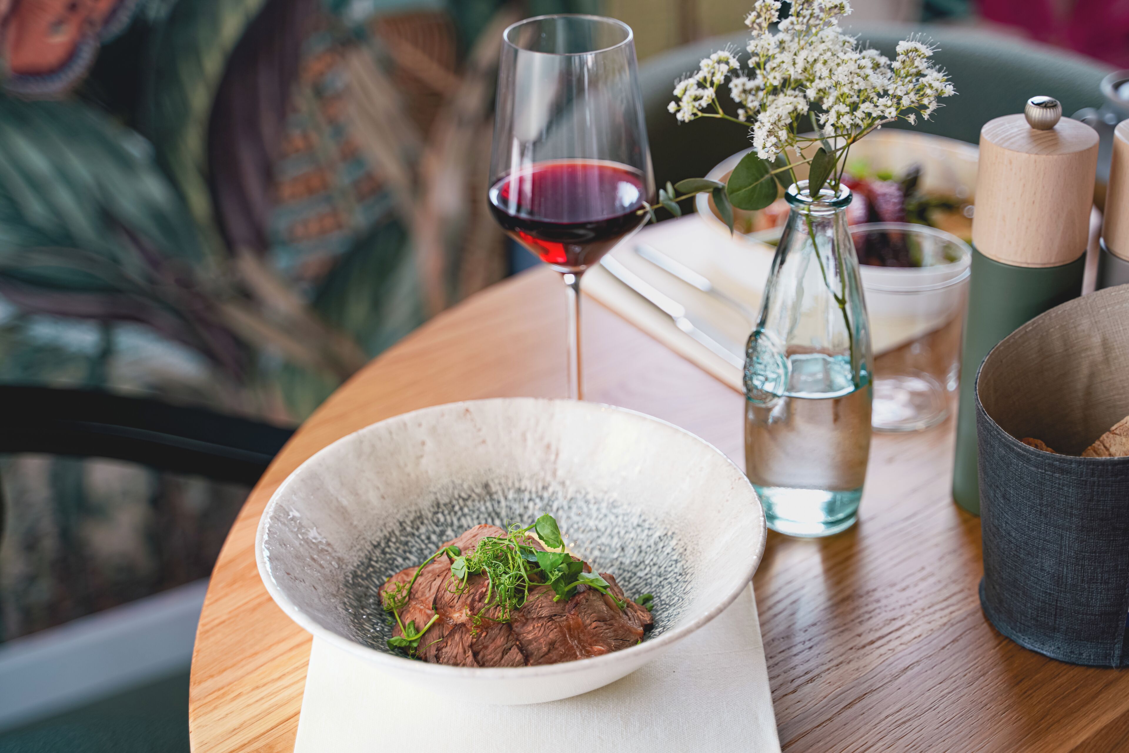 Ein Esstisch mit einer Schüssel Essen, einem Glas Rotwein, einer Wasserflasche und einer Vase mit Blumen.