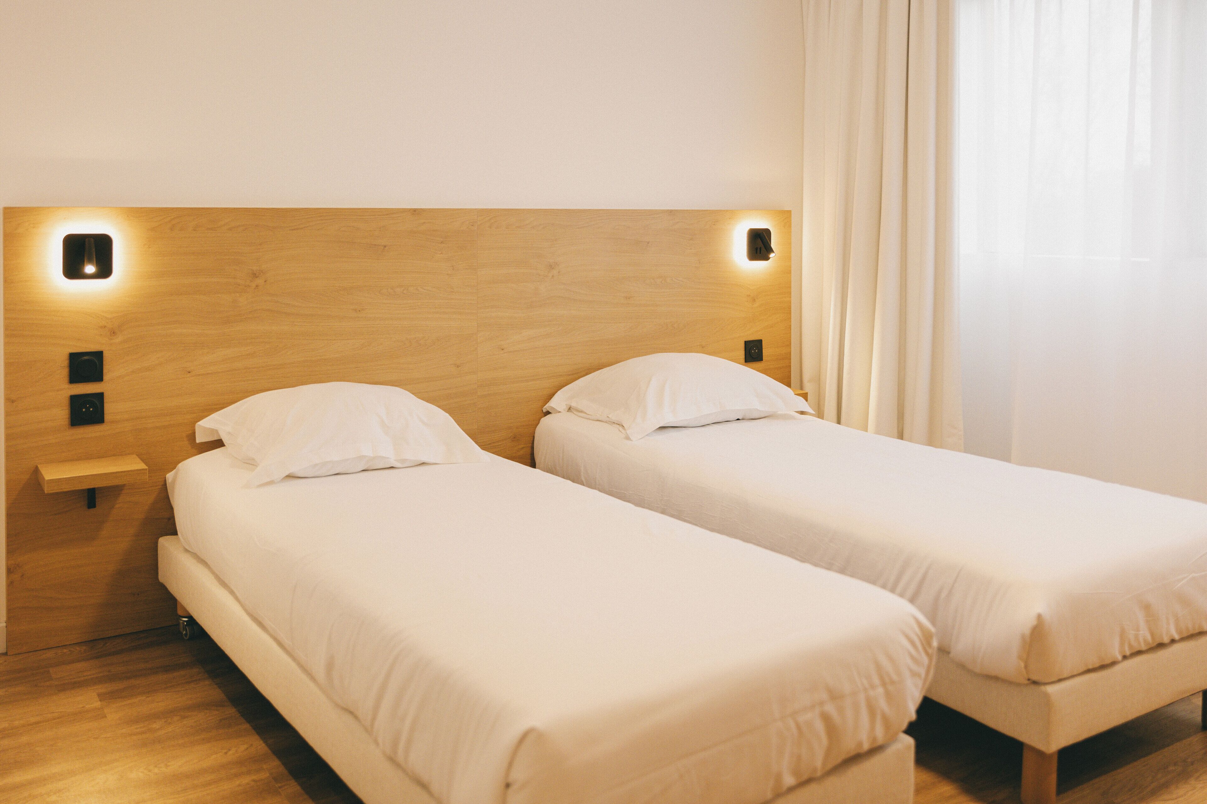 Hotellihuone, jossa on kaksi erillistä sänkyä, puiset päätylevyt, yöpöydät ja ikkuna, jossa on verhot.