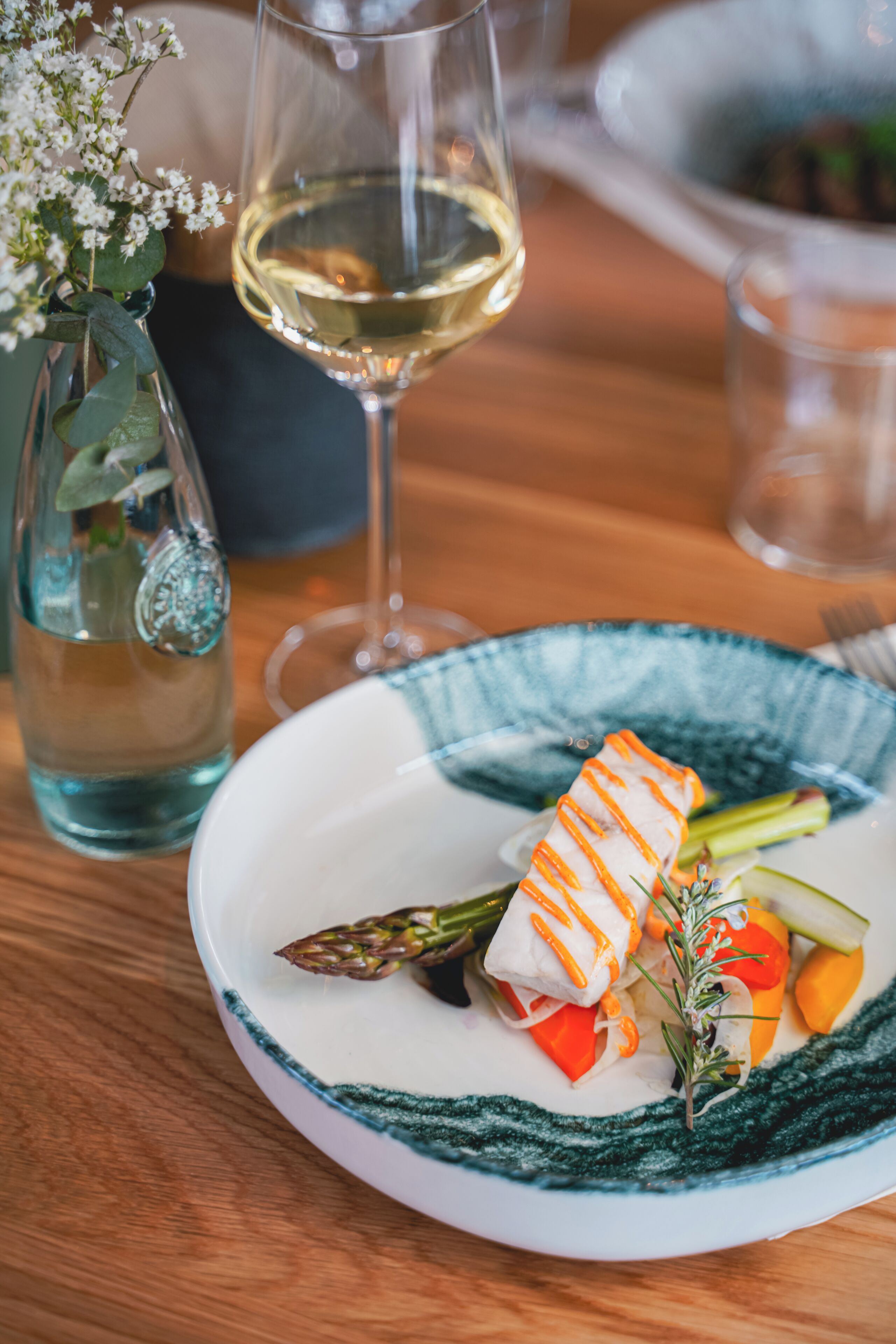 A plate with a rolled appetizer, asparagus, and vegetables on a table with a glass of white wine and a vase with flowers.