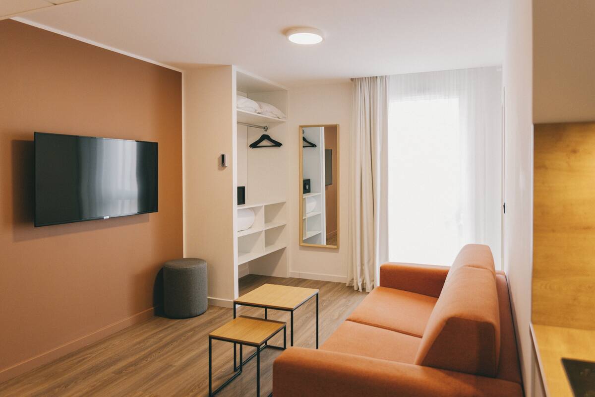 Une chambre d’hôtel moderne équipée d’un téléviseur à écran plat fixé au mur, d’un canapé, d’une petite table en bois, d’un pouf gris et d’une armoire avec des étagères et un miroir.