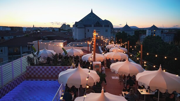 Terraza en la azotea con sombrillas blancas y luces de cuerda, con vistas a un paisaje urbano que incluye edificios y una iglesia.