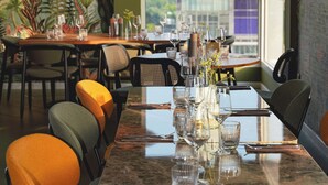 Un restaurante con una larga mesa dispuesta para comer, sillas con cojines naranja y vistas a la ciudad a través de amplias ventanas.