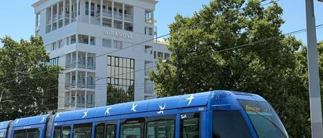A blue tram with passengers, passing by a modern white building with balconies.