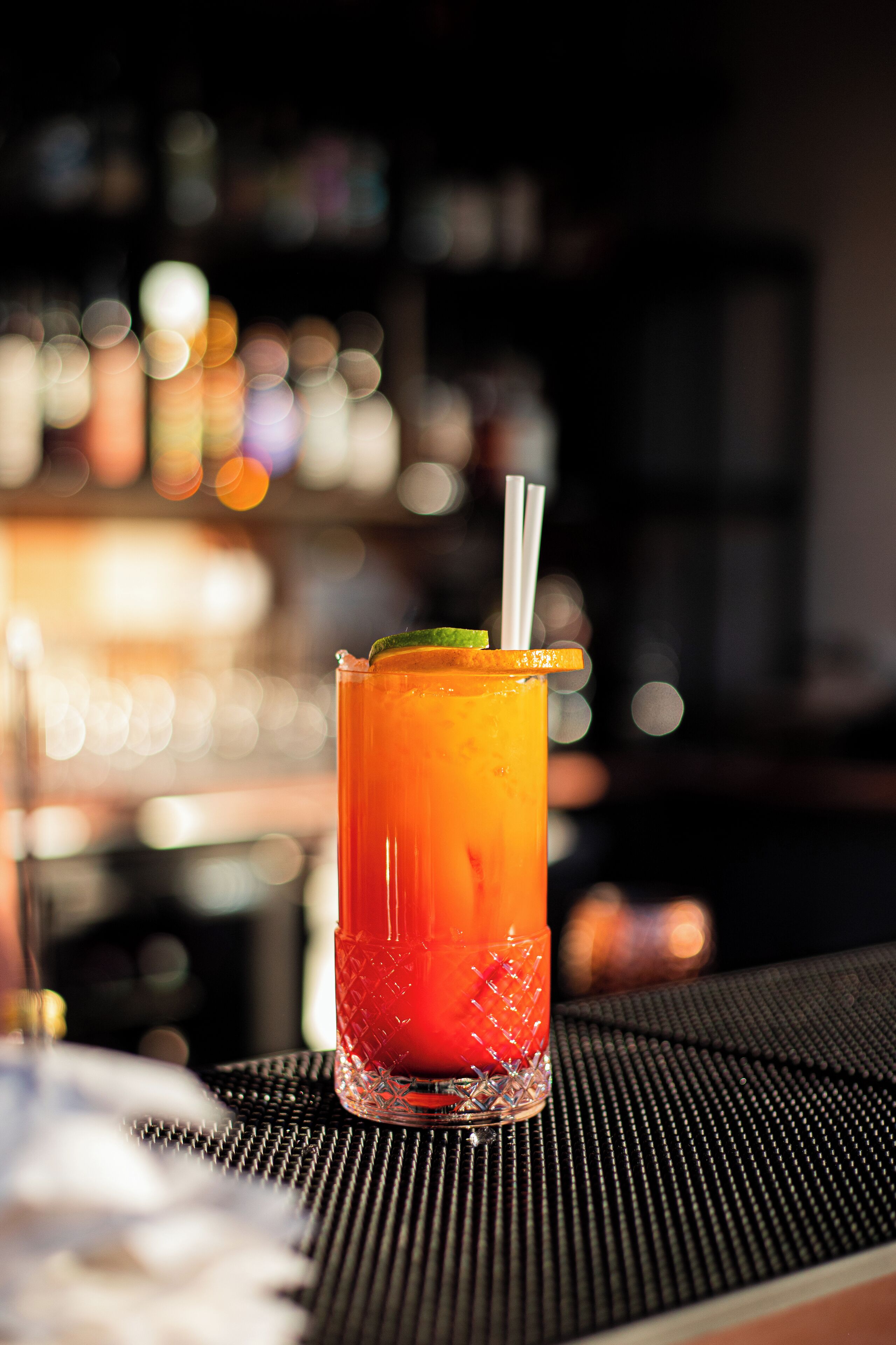 A tall glass with a gradient color, a lime wedge, and a straw, placed on a textured surface.