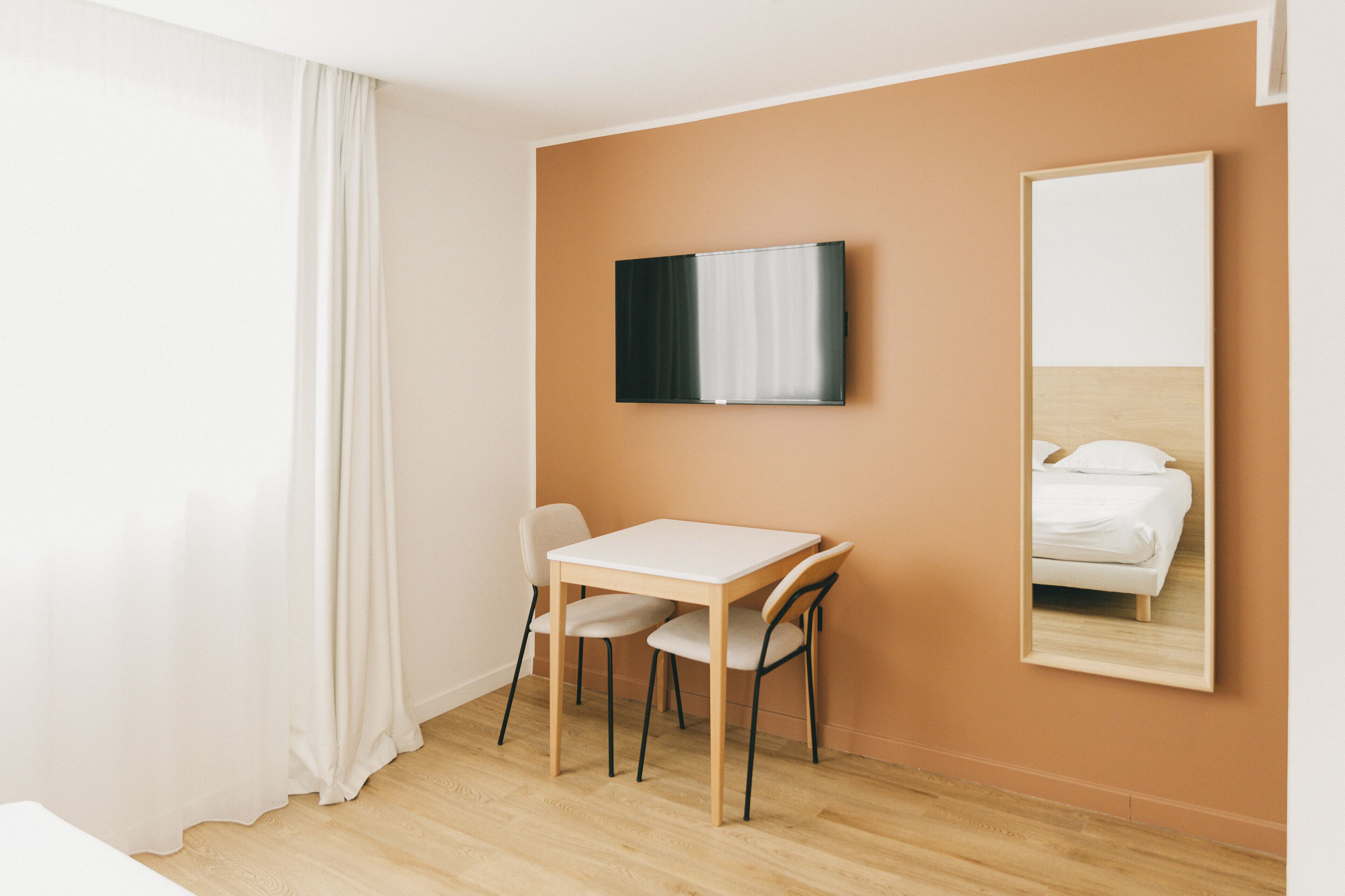 A hotel room with a wooden floor, a white table with two chairs, a flat-screen TV mounted on the wall, and a bed with white bedding.