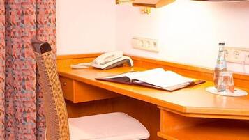 A hotel room with a wooden desk, a telephone, an open book, a bottle of water, and a small stool.