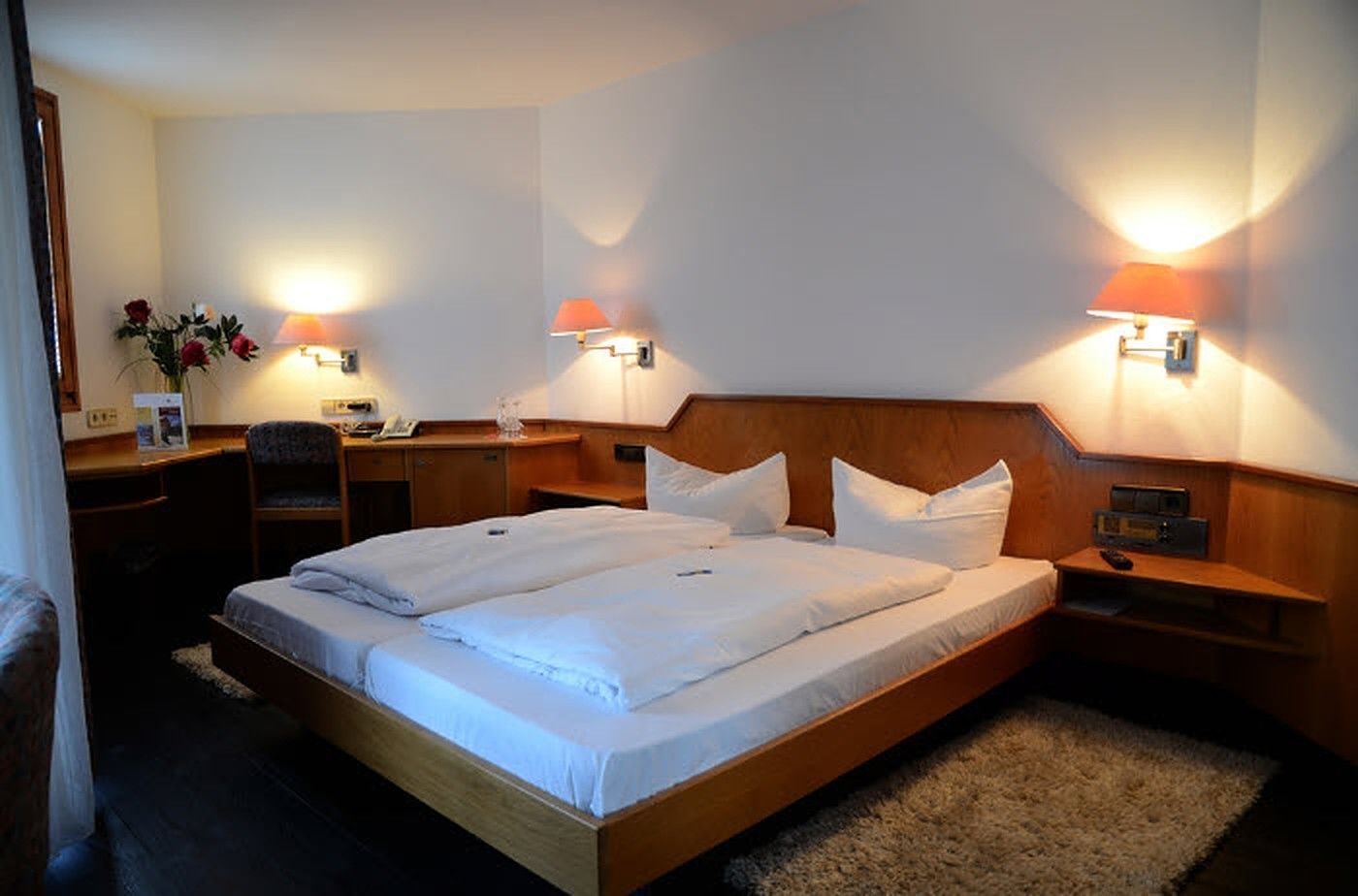 A hotel room with a large bed, two bedside tables, a desk, and two wall-mounted lamps.