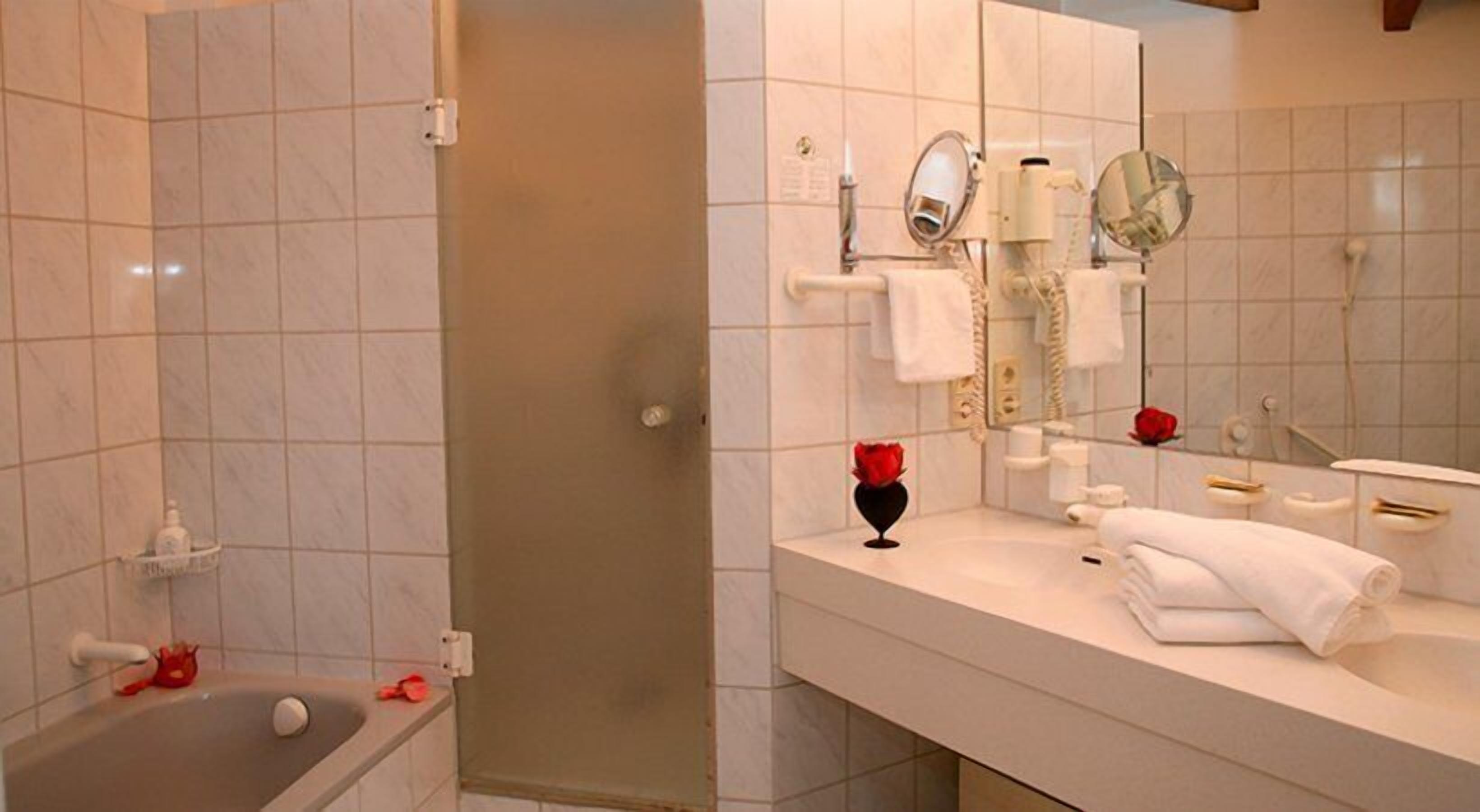 Un baño con bañera, ducha, tocador con espejo y un jarrón con una rosa roja.