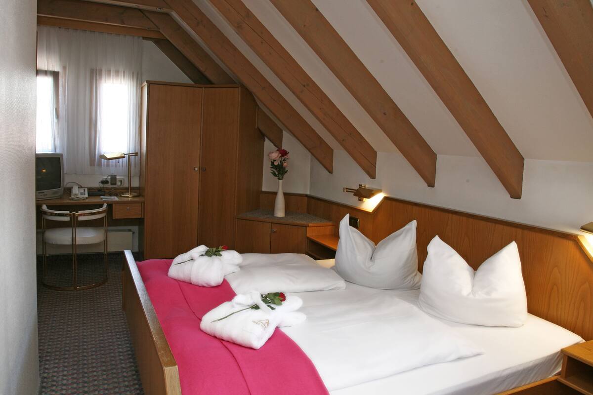 A hotel room with a wooden ceiling, a bed with white linens and pillows, a desk with a chair, and a small table with a vase of flowers.