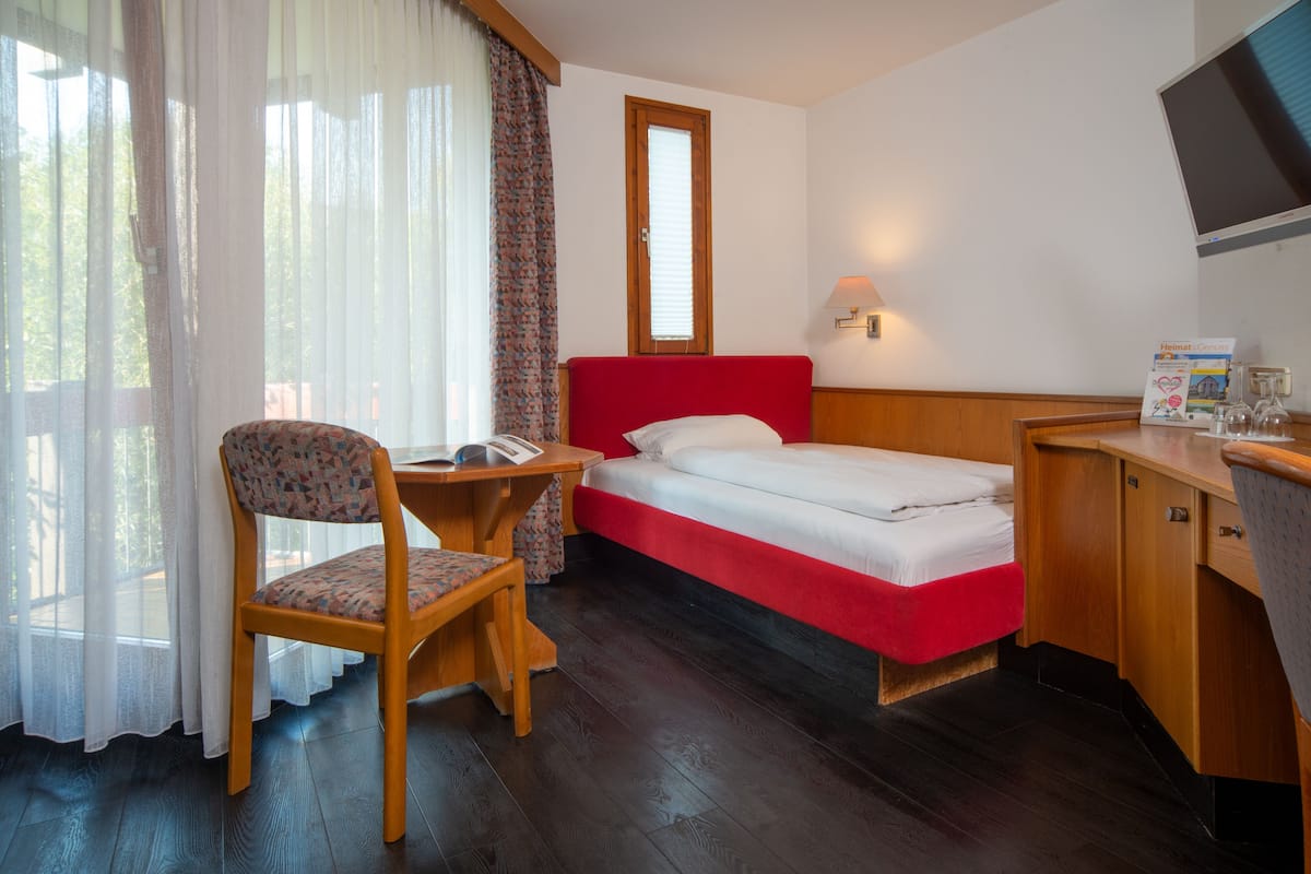A compact hotel room with a single bed, a wooden desk, a chair, a small table, a TV, and a window with curtains.
