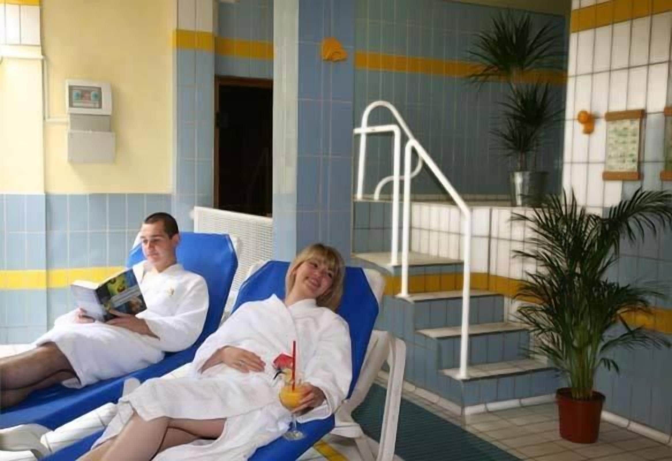 Due persone si rilassano su lettini in una stanza con piastrelle, piscina, una scala e piante in vaso.