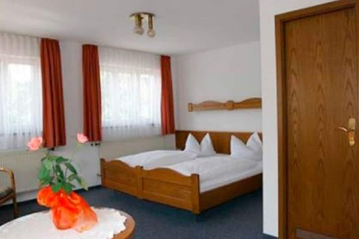 Un dormitorio con una cama de madera, ropa de cama blanca, una mesa redonda con un florero y una puerta de madera.