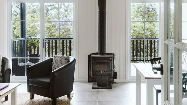 A living room with a black sofa, a white coffee table, and a fireplace. There are large windows with a view of trees and a balcony with a railing. - Possums Retreat - Private Spacious Cottage with Hydrotherapy Spa (North Maleny)