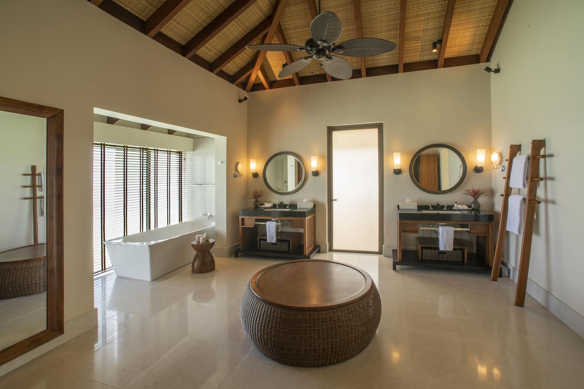 A spacious bathroom with a large round wicker ottoman, two sinks with mirrors, and a ceiling fan.