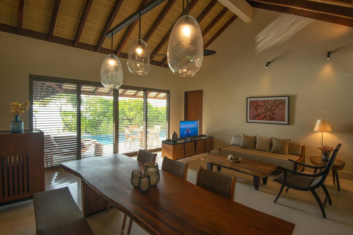 A spacious living area with a wooden dining table, comfortable seating, a television, and a large window with blinds.