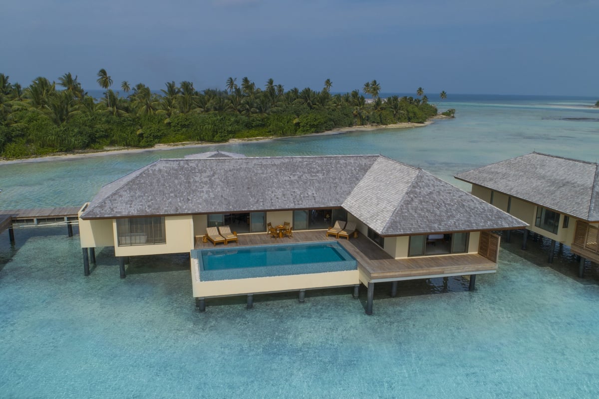 An overwater bungalow with a pool, surrounded by clear turquoise water and palm trees.