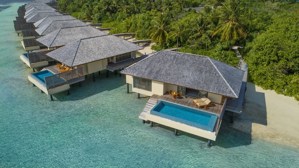 An aerial view of overwater bungalows with private pools, surrounded by lush greenery and clear turquoise waters.