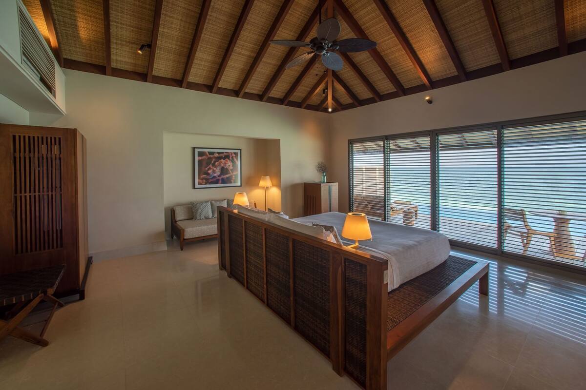 A bedroom with a bed, bedside lamps, a sofa, a painting, a ceiling fan, and large windows.
