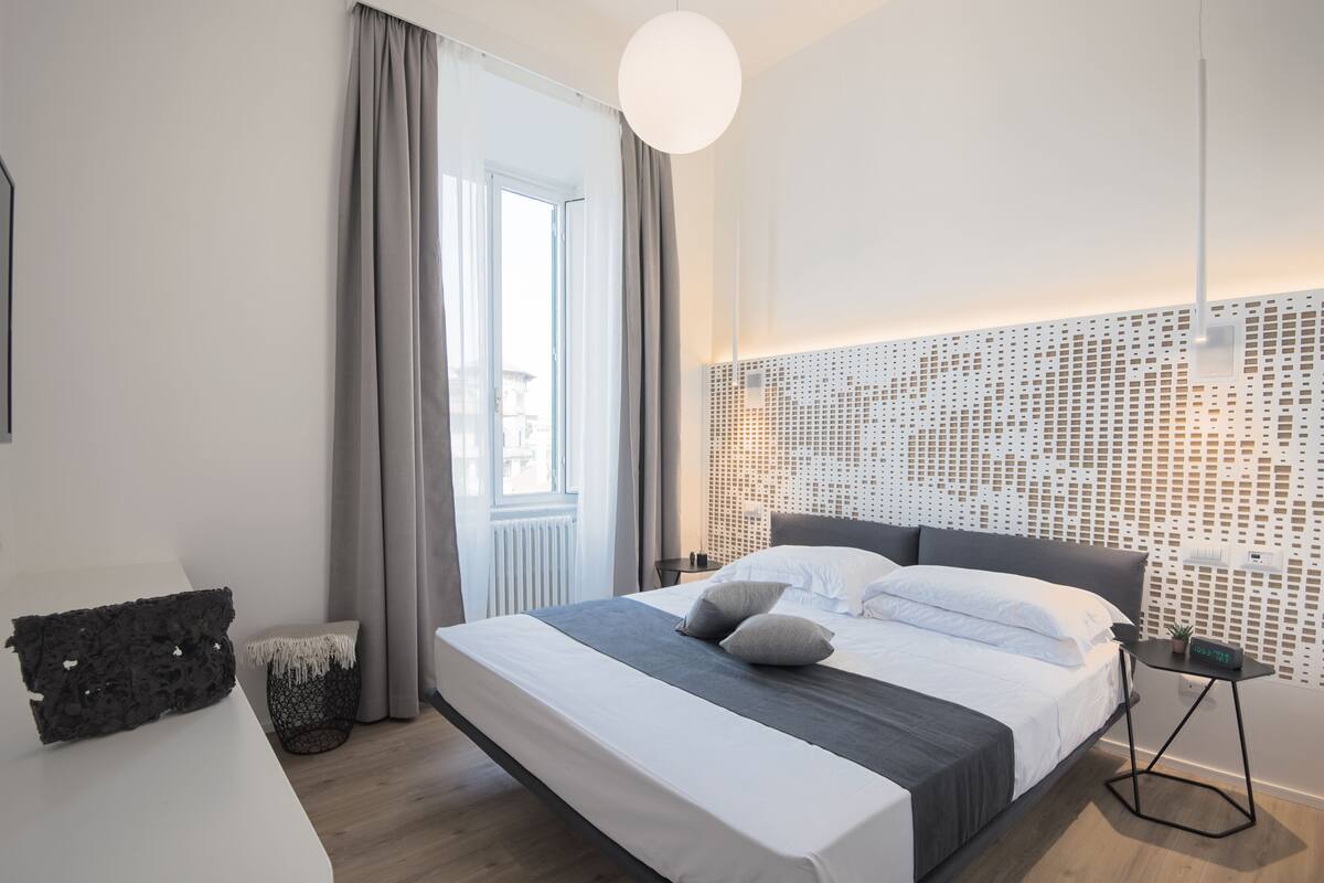 A modern hotel room with a large bed, a bedside table, a small side table, and a decorative wall panel.