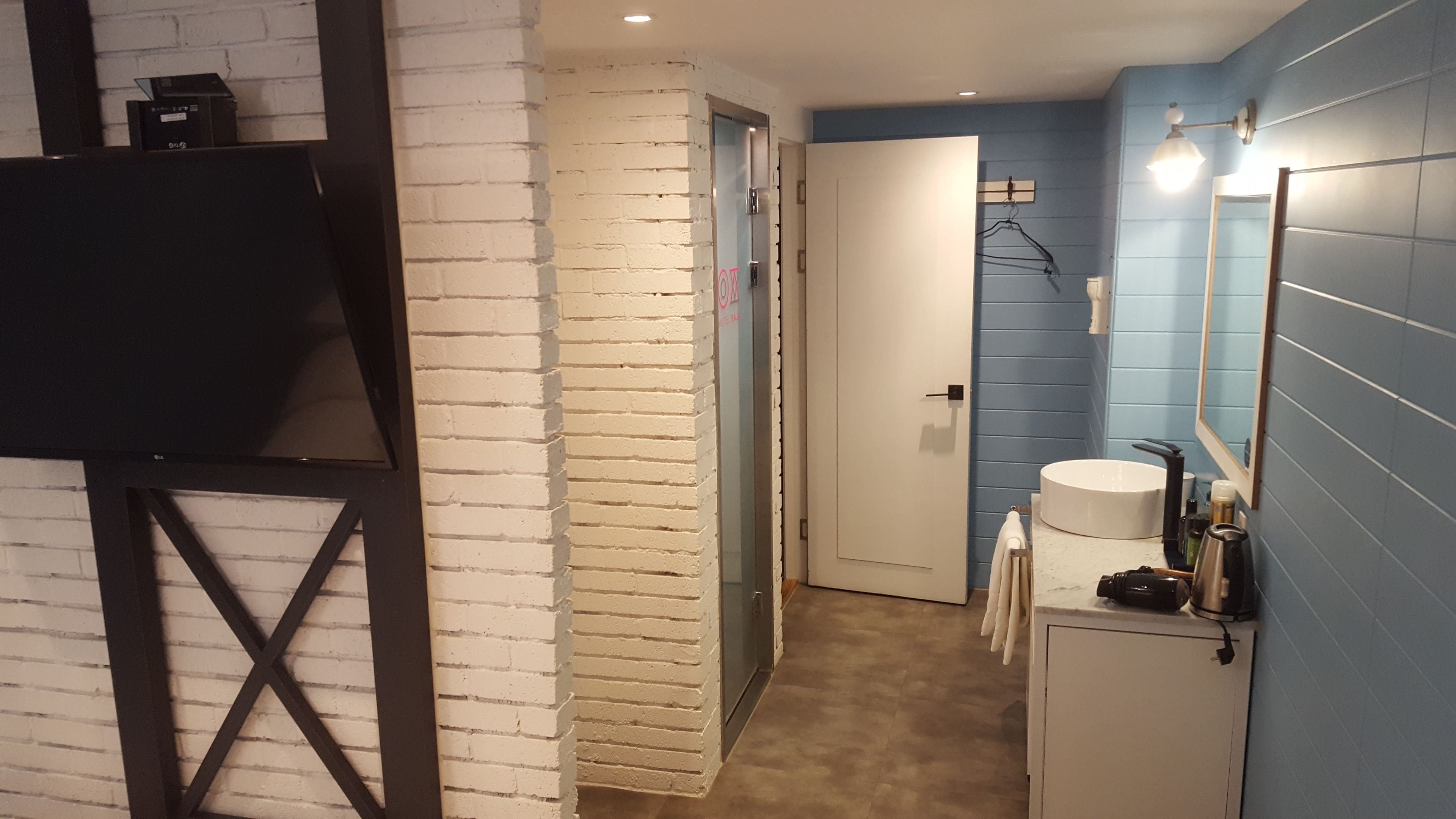 Un estrecho pasillo con pared de ladrillo blanco, una puerta, un mueble blanco con lavamanos y un espejo.