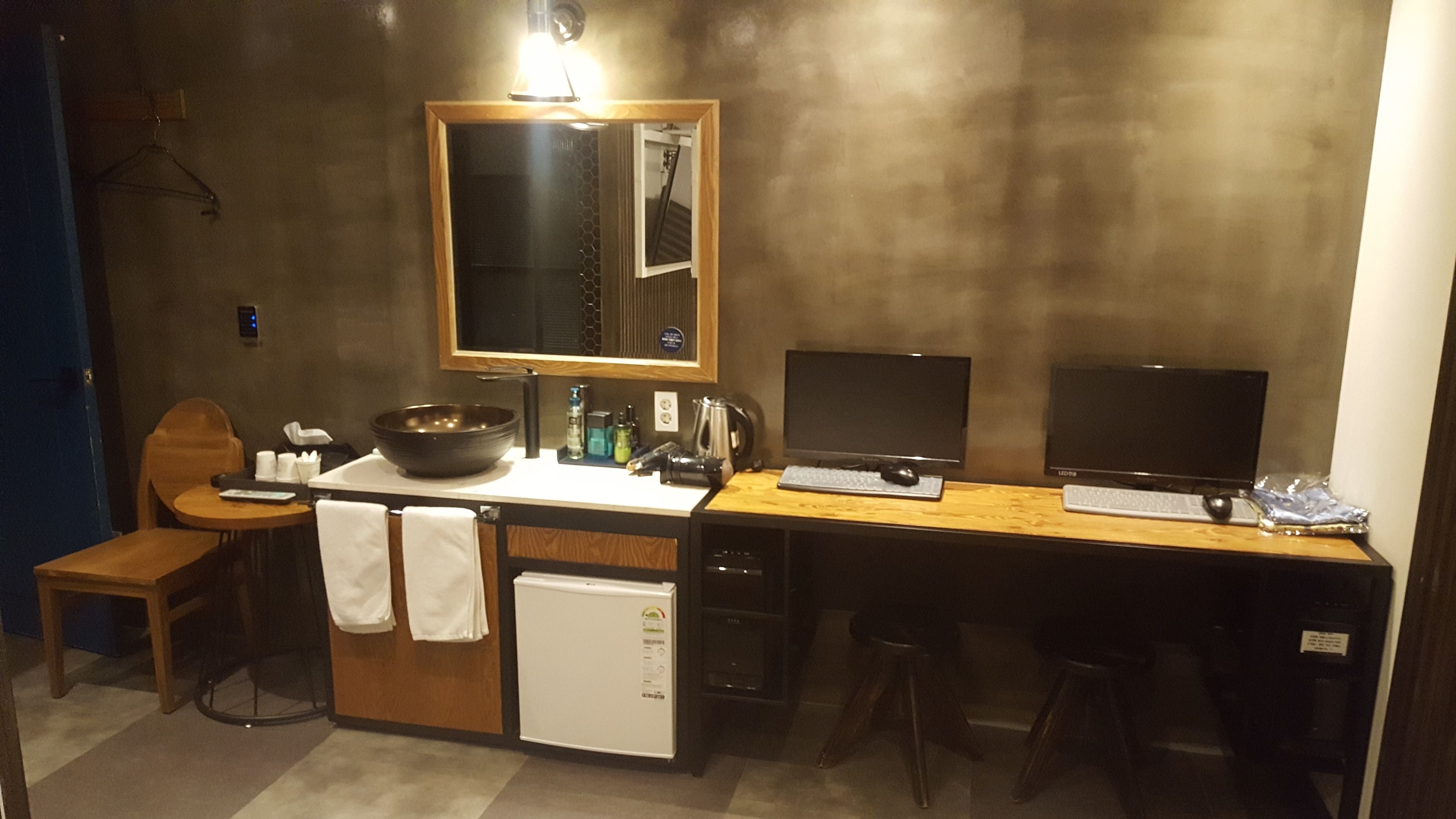 A compact hotel room with a kitchenette, a single bed, a small bathroom, and a desk with two monitors.