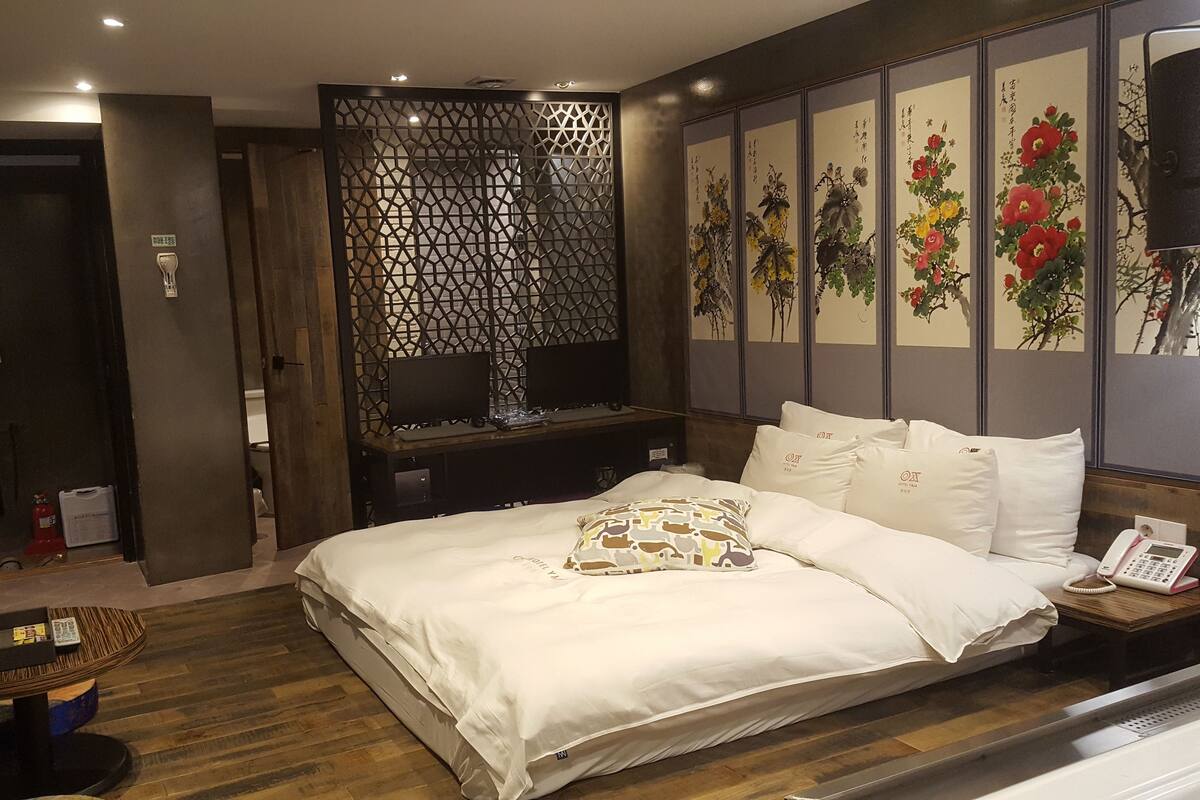 Une chambre d’hôtel avec un grand lit, un bureau équipé de deux ordinateurs, un paravent décoratif et un mur orné d’œuvres d’art florales.