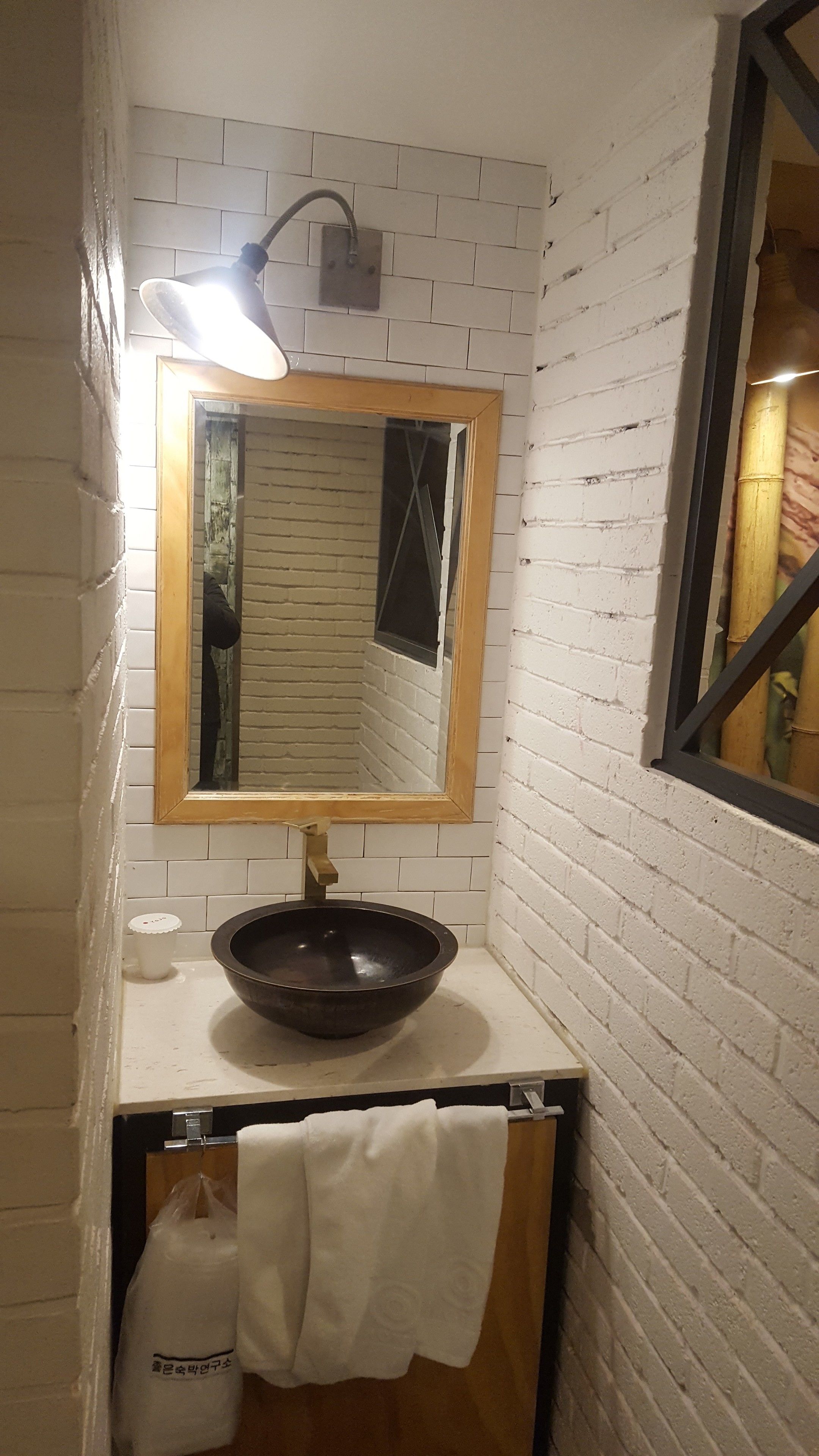 A bathroom with a white brick wall, a wooden vanity, a black basin, a mirror with a wooden frame, a towel hanging on the wall, and a small window with a bamboo curtain.