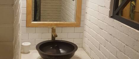 A bathroom with a white brick wall, a wooden vanity, a black basin, a mirror with a wooden frame, a towel hanging on the wall, and a small window with a bamboo curtain.