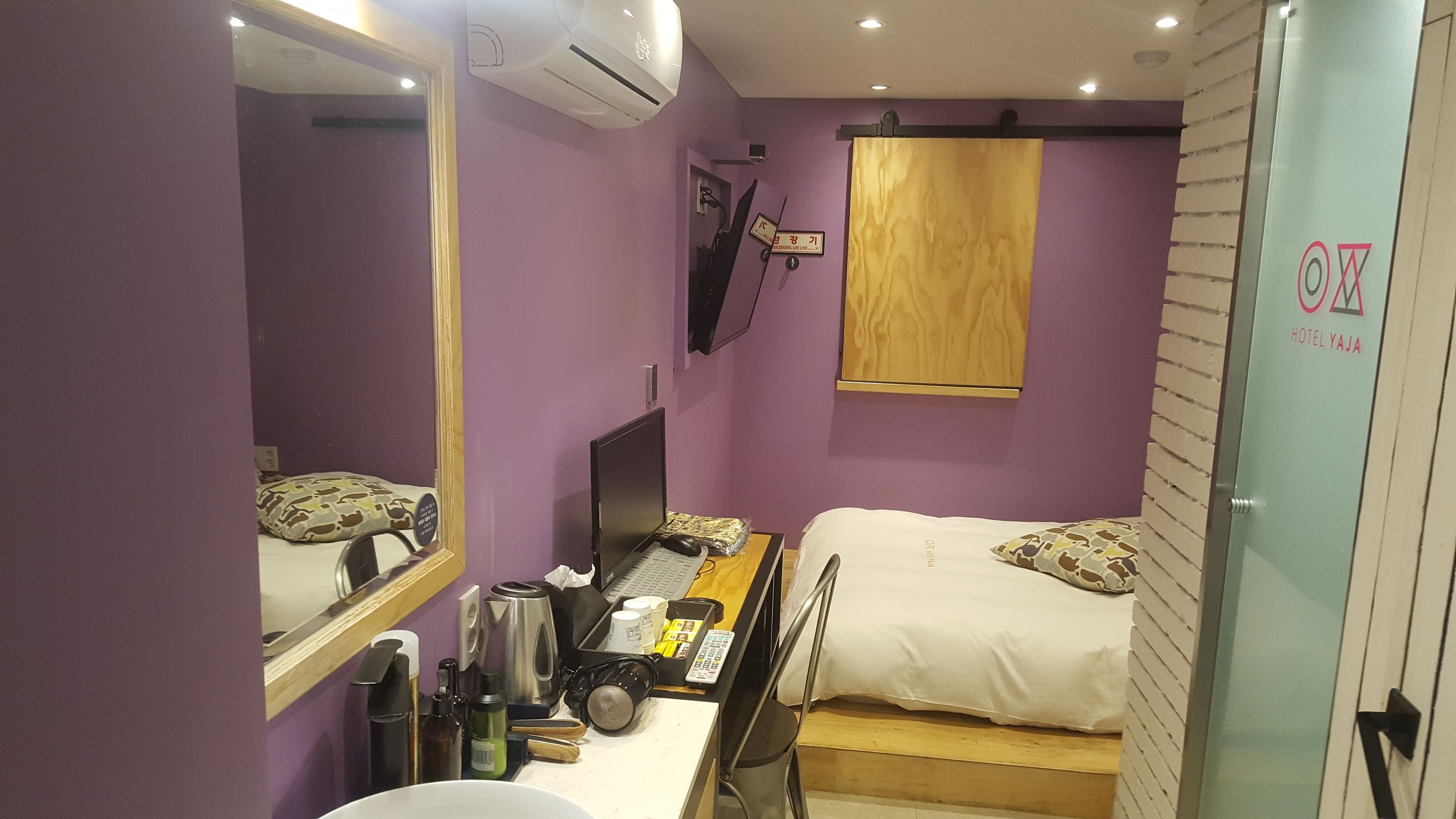 Une chambre d’hôtel compacte aux murs violets, avec un lit agrémenté d’oreillers à motifs, un bureau avec un ordinateur, un miroir et un téléviseur fixé au mur.