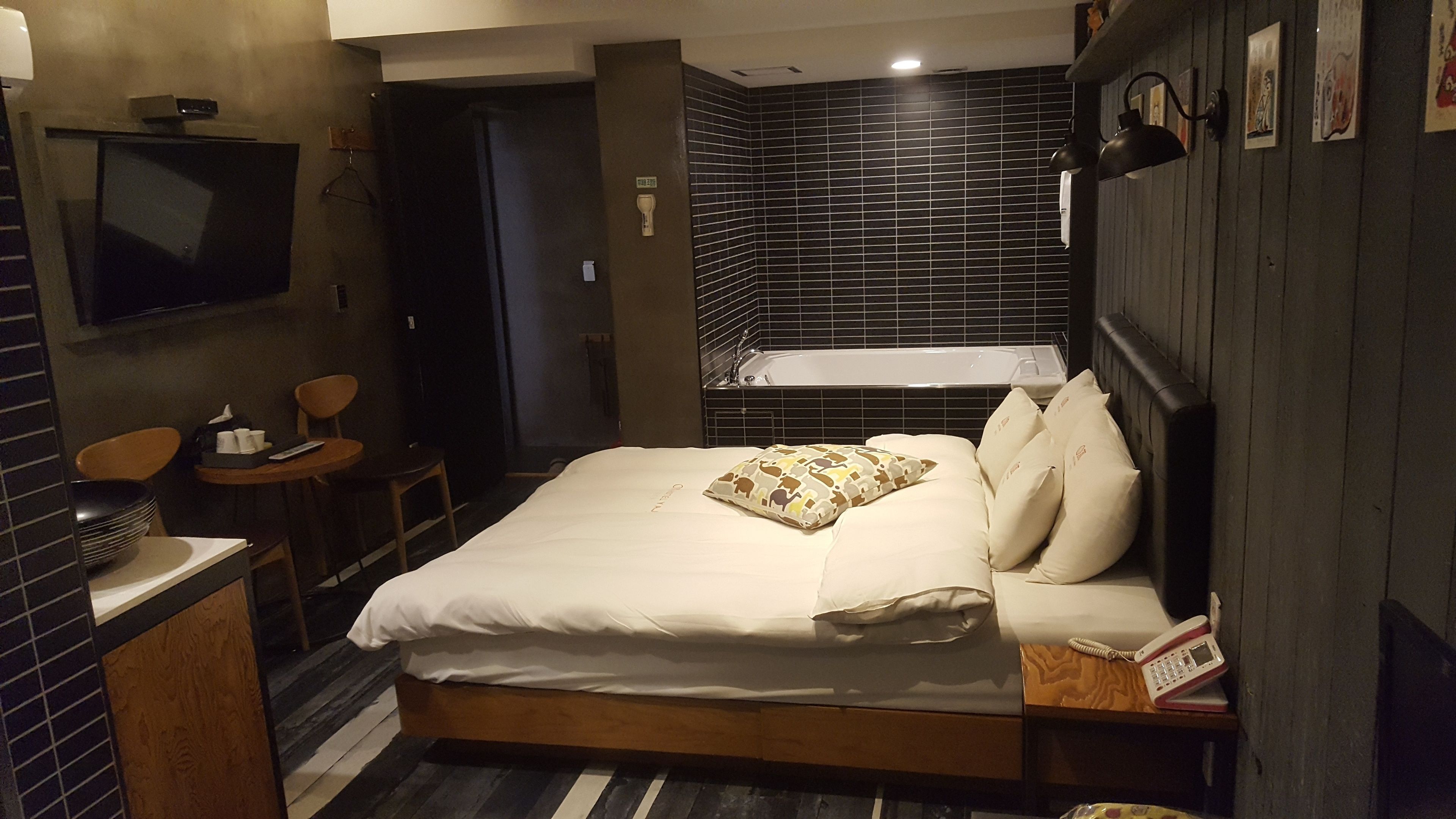 A hotel room with a bed, a bedside table, a chair, a small table with a remote control, a wall-mounted TV, and a bathtub.