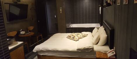 A hotel room with a bed, a bedside table, a chair, a small table with a remote control, a wall-mounted TV, and a bathtub.