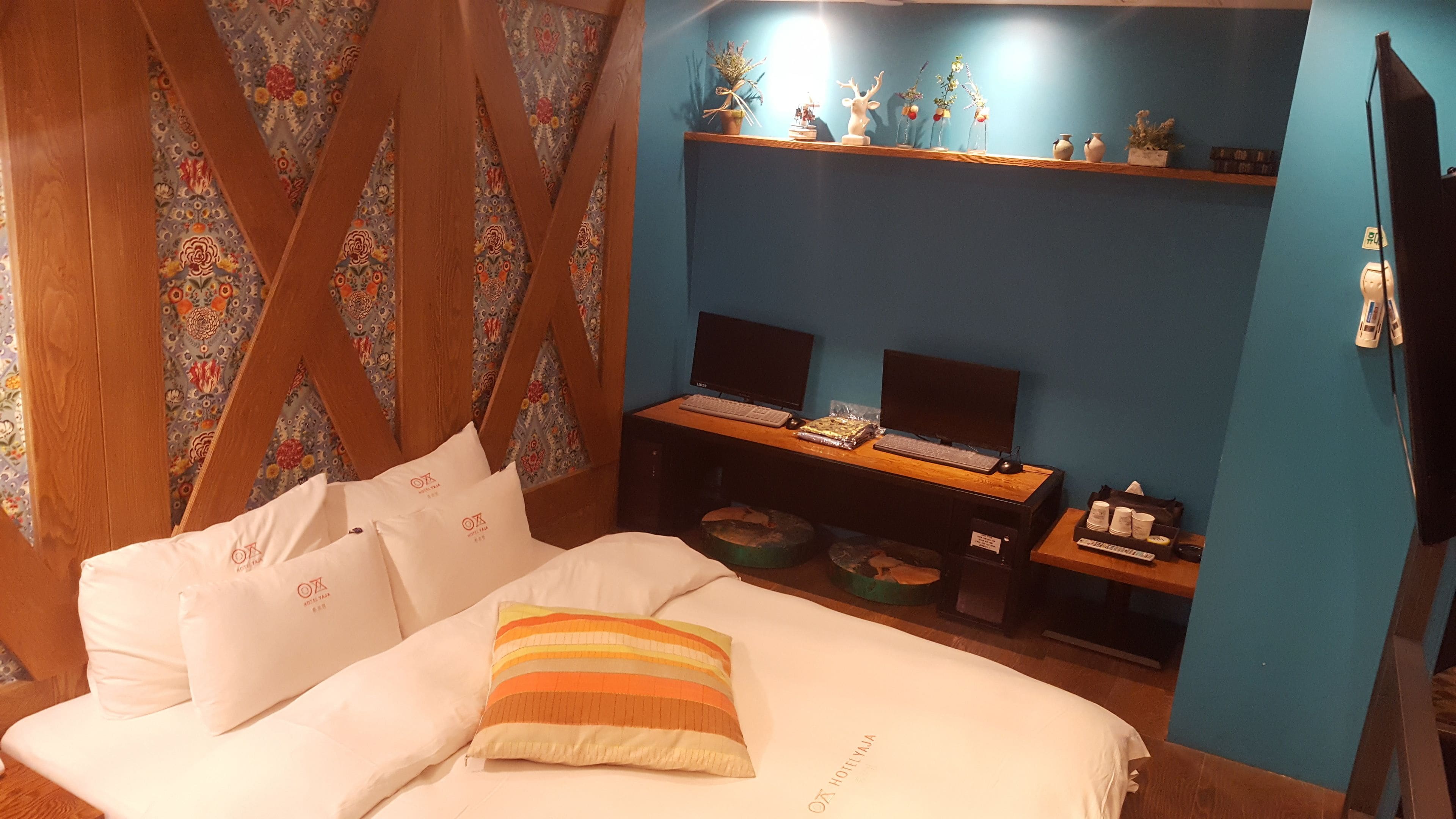 Una habitación de hotel con una cama, un escritorio con dos monitores, un estante con adornos y una pared azul de acento.
