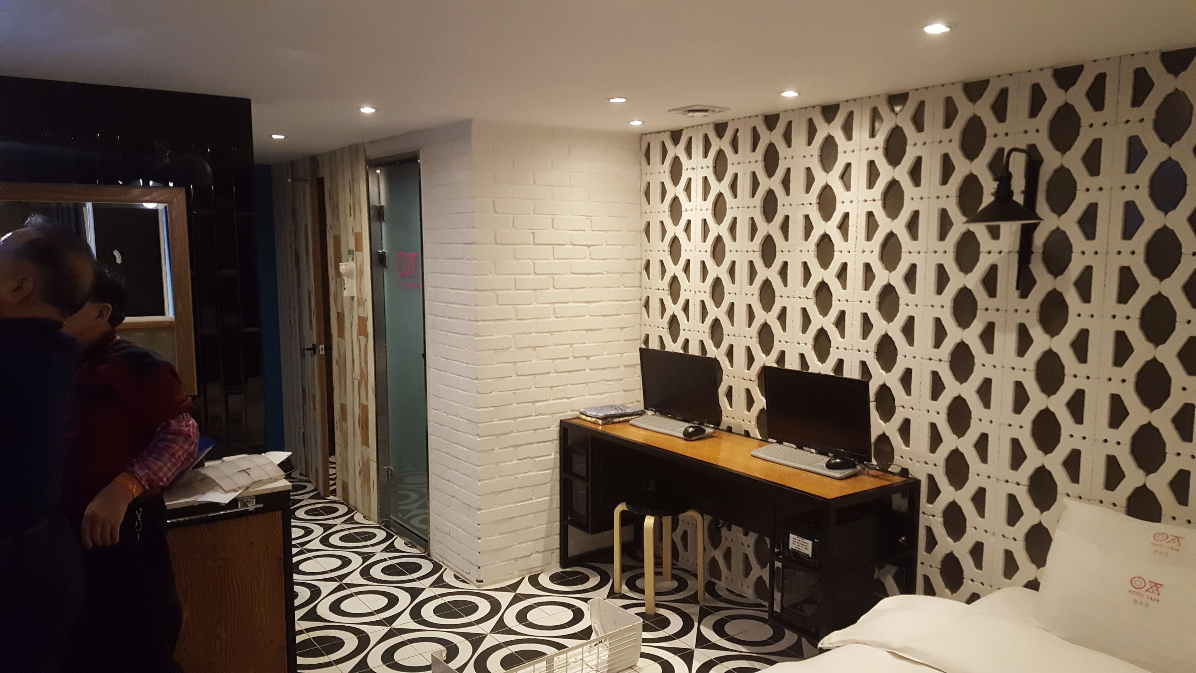 A hotel room with a bed, a desk with two monitors, a patterned wall, and a door leading to another room.