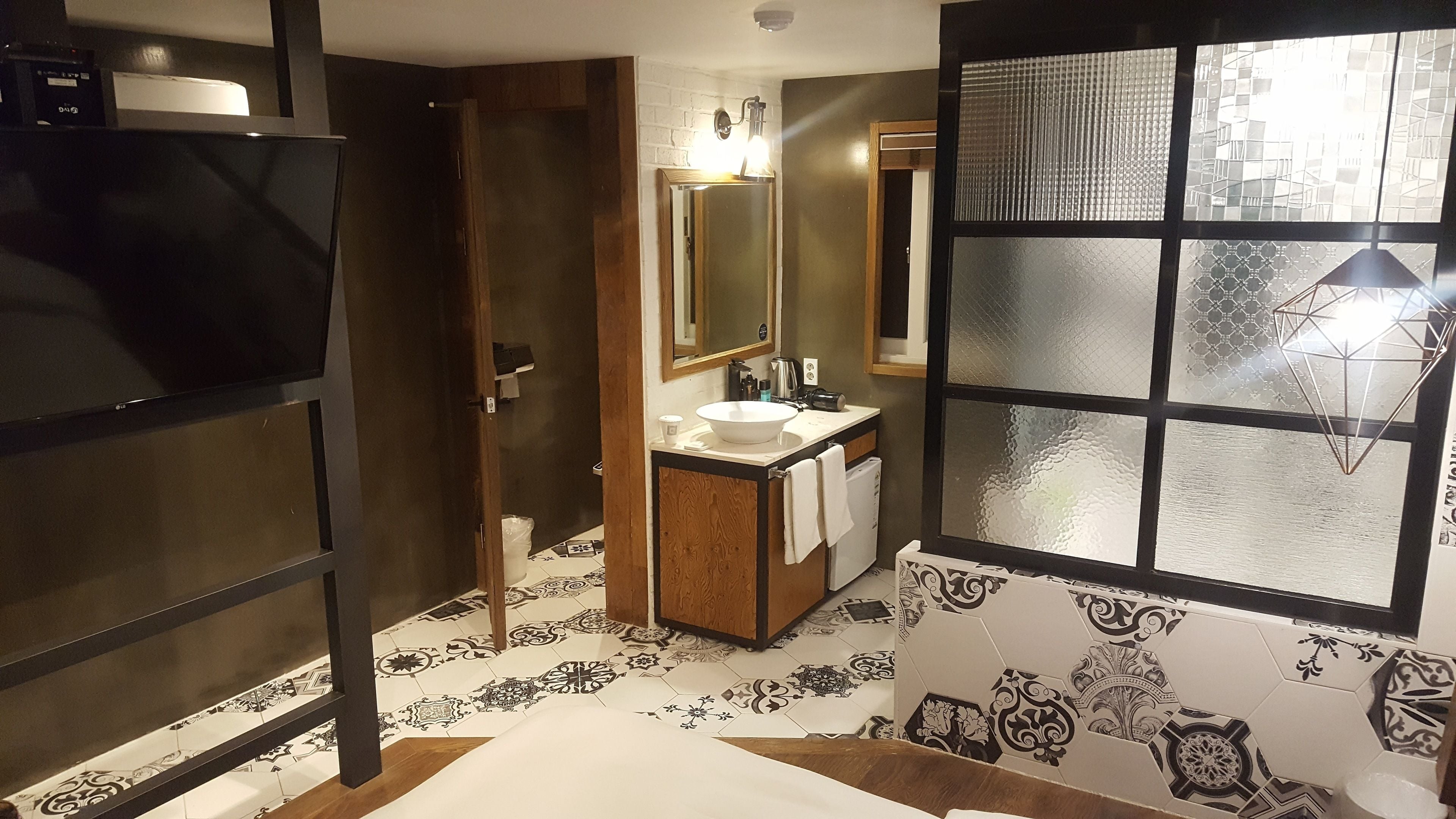 Une salle de bain avec un grand miroir, un lavabo et des toilettes. Il y a une fenêtre ornée d’un motif géométrique.