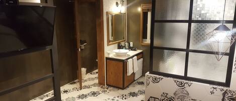 Une salle de bain avec un grand miroir, un lavabo et des toilettes. Il y a une fenêtre ornée d’un motif géométrique.