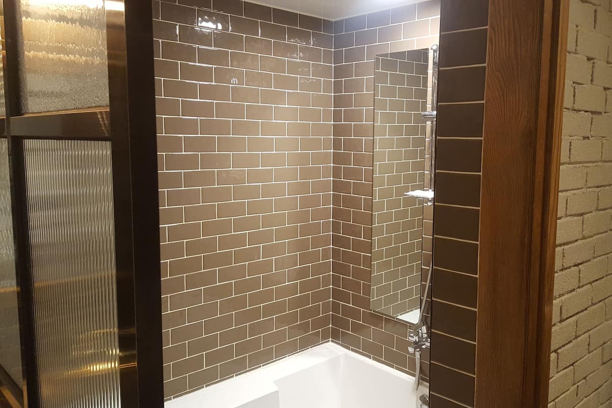 Une salle de bain d’angle avec une baignoire blanche et un espace douche, le tout entouré de murs recouverts de carreaux bruns.