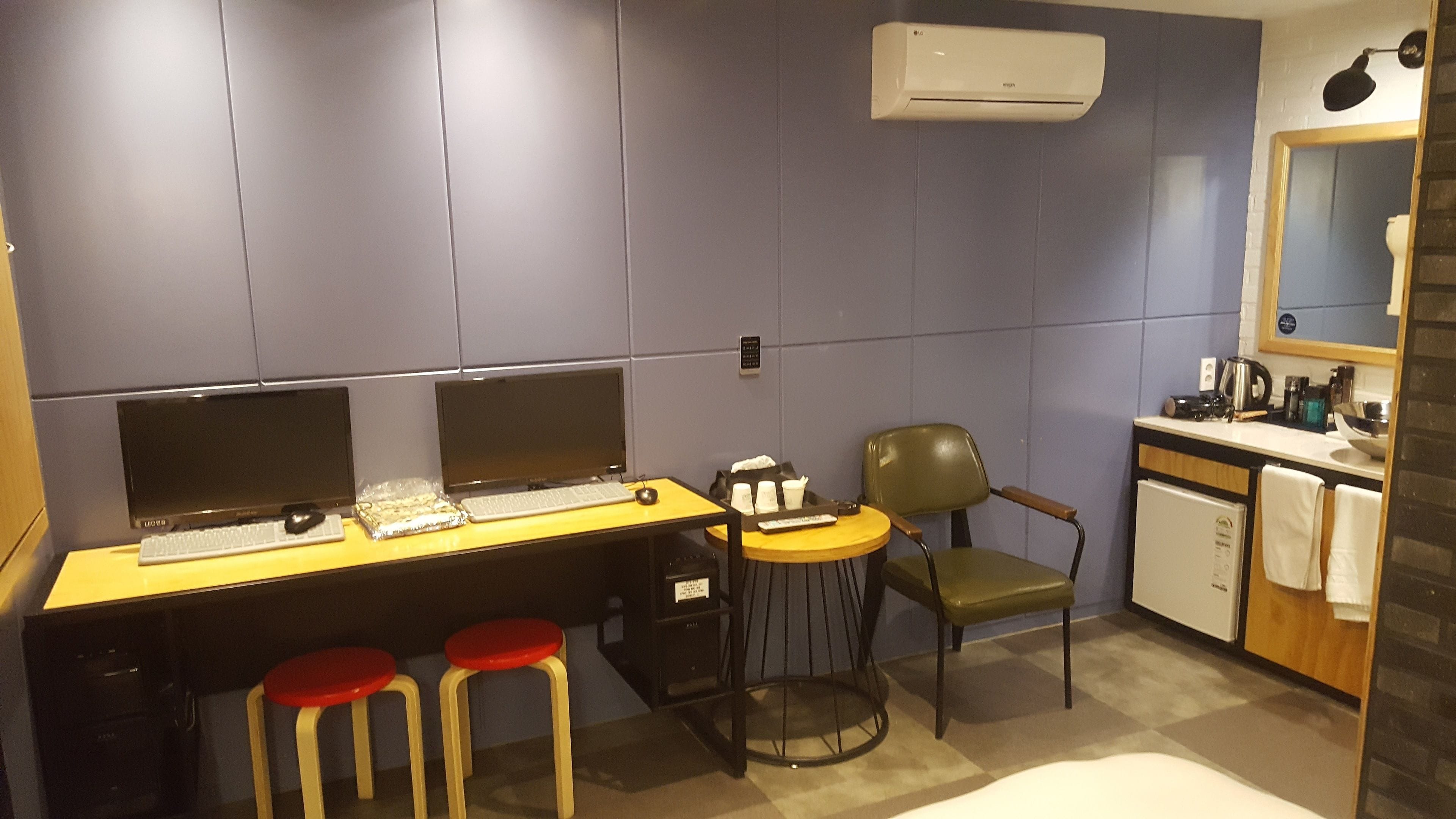 A modern office space with two desks, each with a computer and keyboard, and two red stools. There is a chair, a small table with a bowl, and a coffee machine.