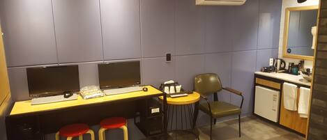 A modern office space with two desks, each with a computer and keyboard, and two red stools. There is a chair, a small table with a bowl, and a coffee machine.