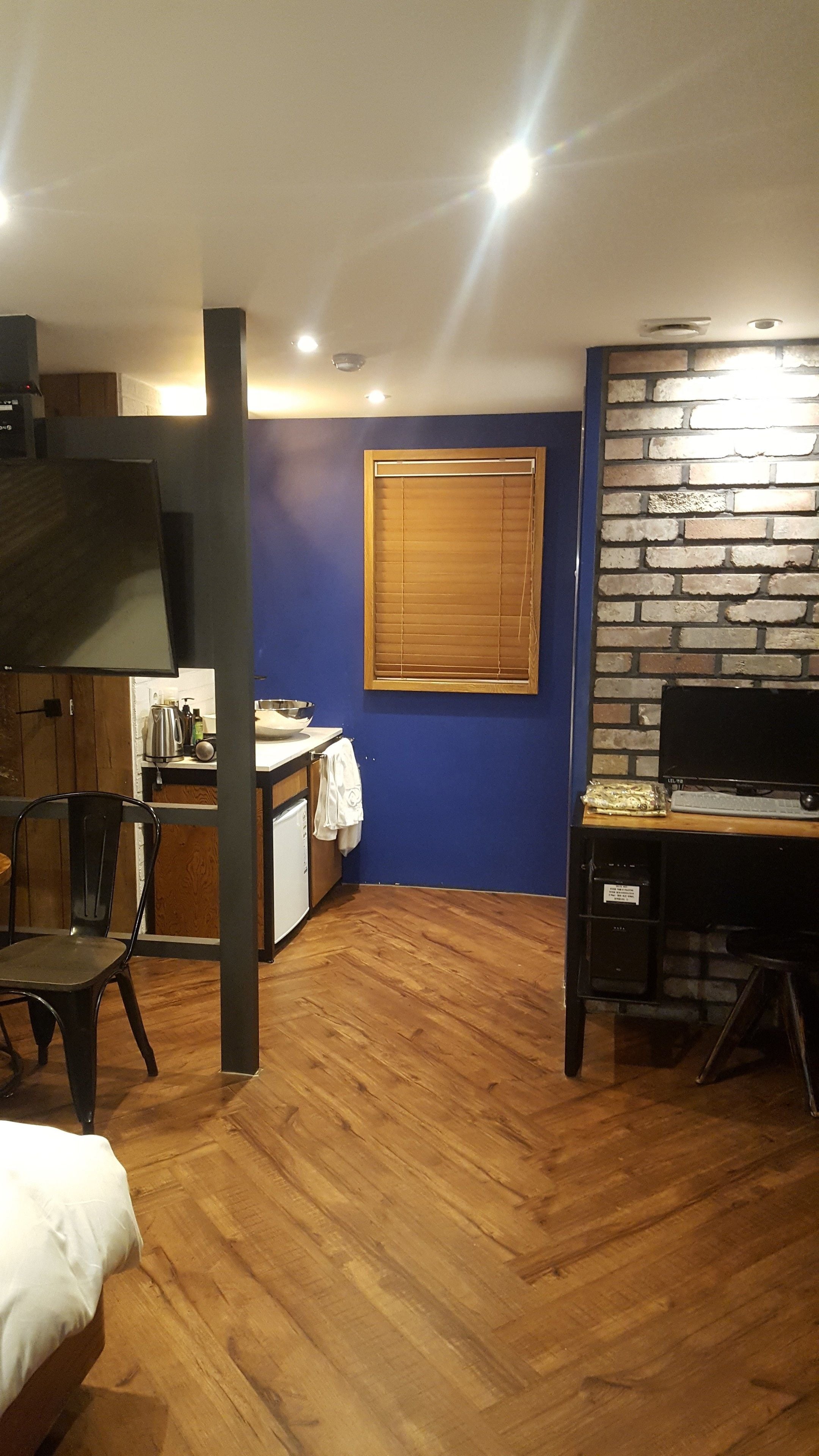 Une pièce de taille modeste avec un plancher en bois, un coin cuisine, un téléviseur, un mur en briques et une fenêtre avec des stores.