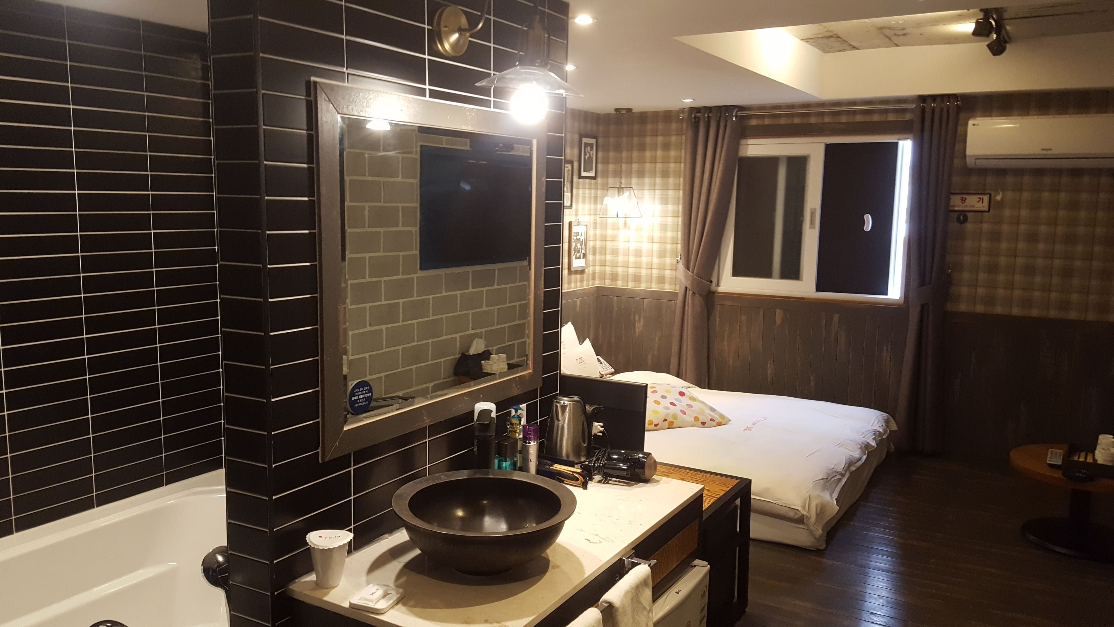 Une chambre d’hôtel avec un mur recouvert de carreaux noirs, un grand miroir, un lit avec une couette blanche, un plancher en bois et une fenêtre avec des rideaux.
