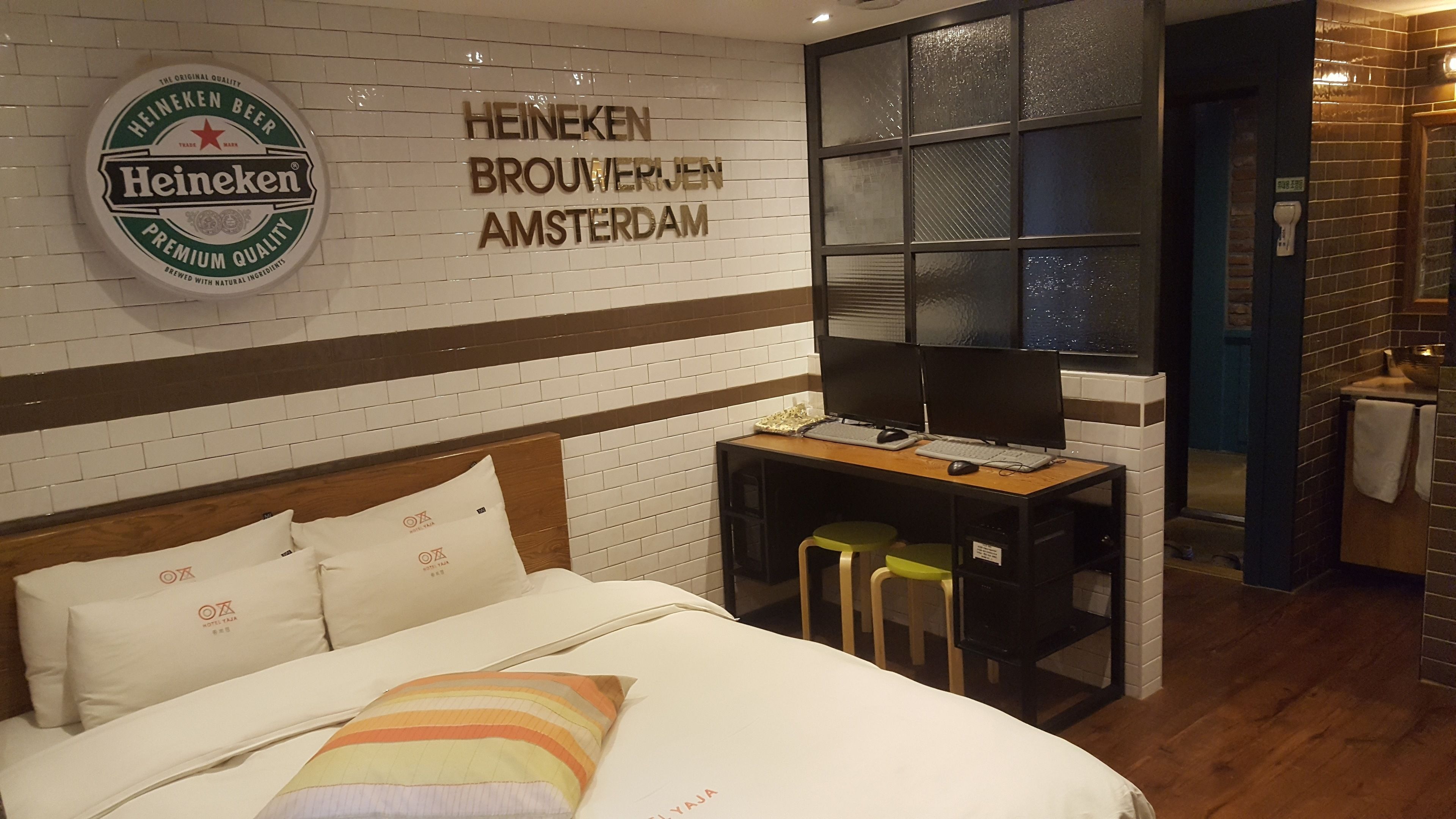 A hotel room with a bed, a desk with a computer, and a Heineken sign on the wall.