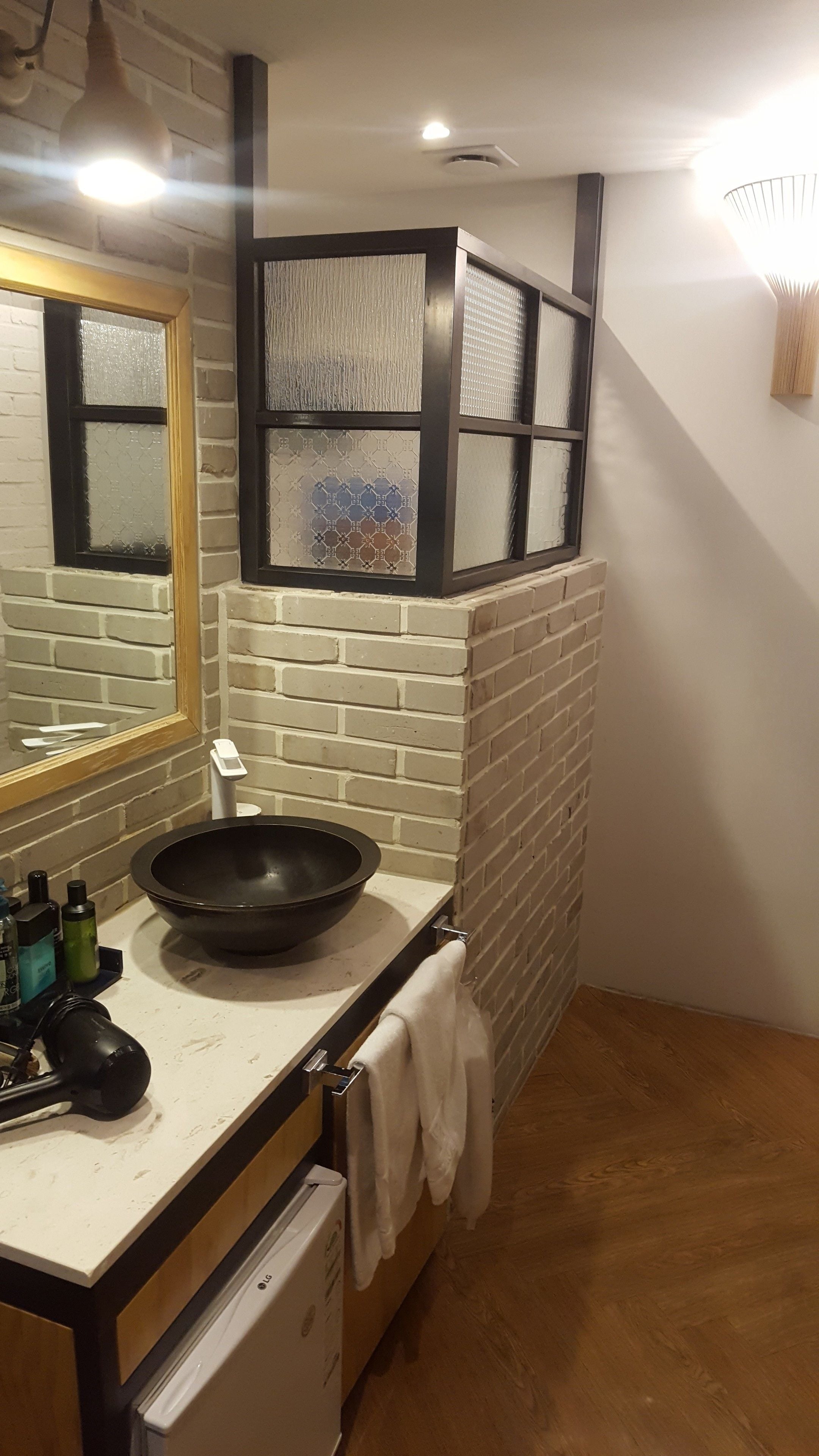 A kitchen with a white countertop, a black frying pan, a sink, and a mirror. There is a towel hanging on the wall and a white appliance below the countertop.