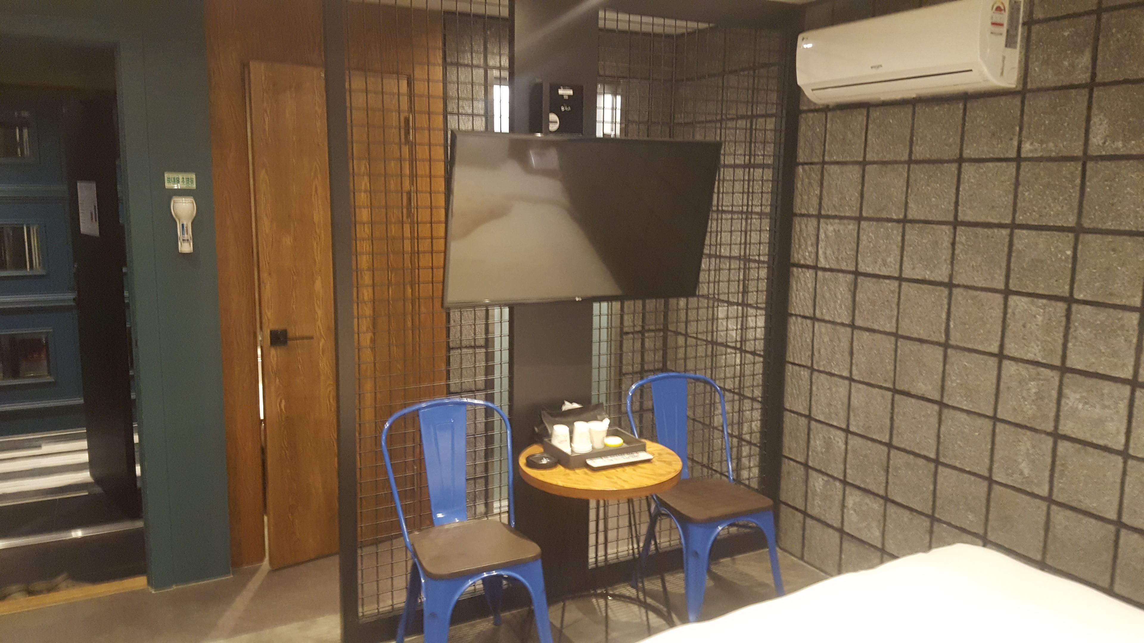 A room with a bed, a TV mounted on the wall, two blue chairs, a small table, and an air conditioning unit.